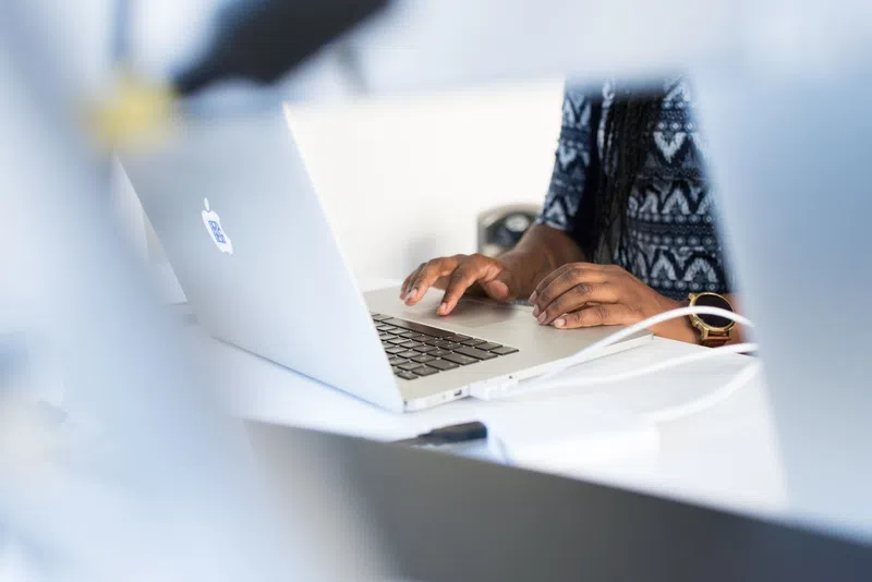 Person typing on a silver MacBook laptop at a white desk with charging cables connected.