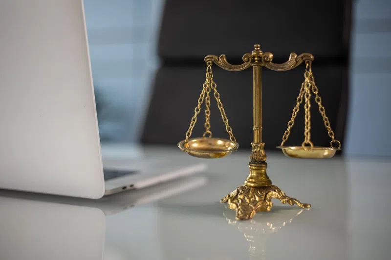 Golden balance scale on glass desk next to open laptop in modern office setting with blurred background.