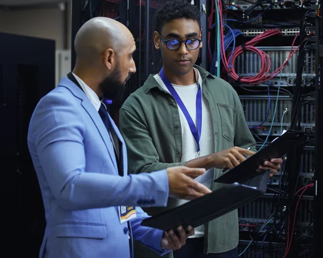 Two IT professionals discussing data on tablets in a server room filled with networking equipment.