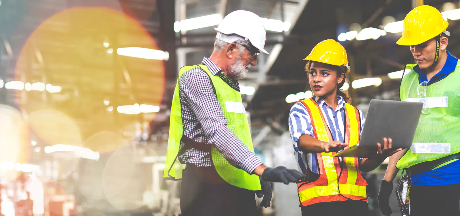 Three workers in hard hats and vests discussing plans with a laptop in a warehouse setting.