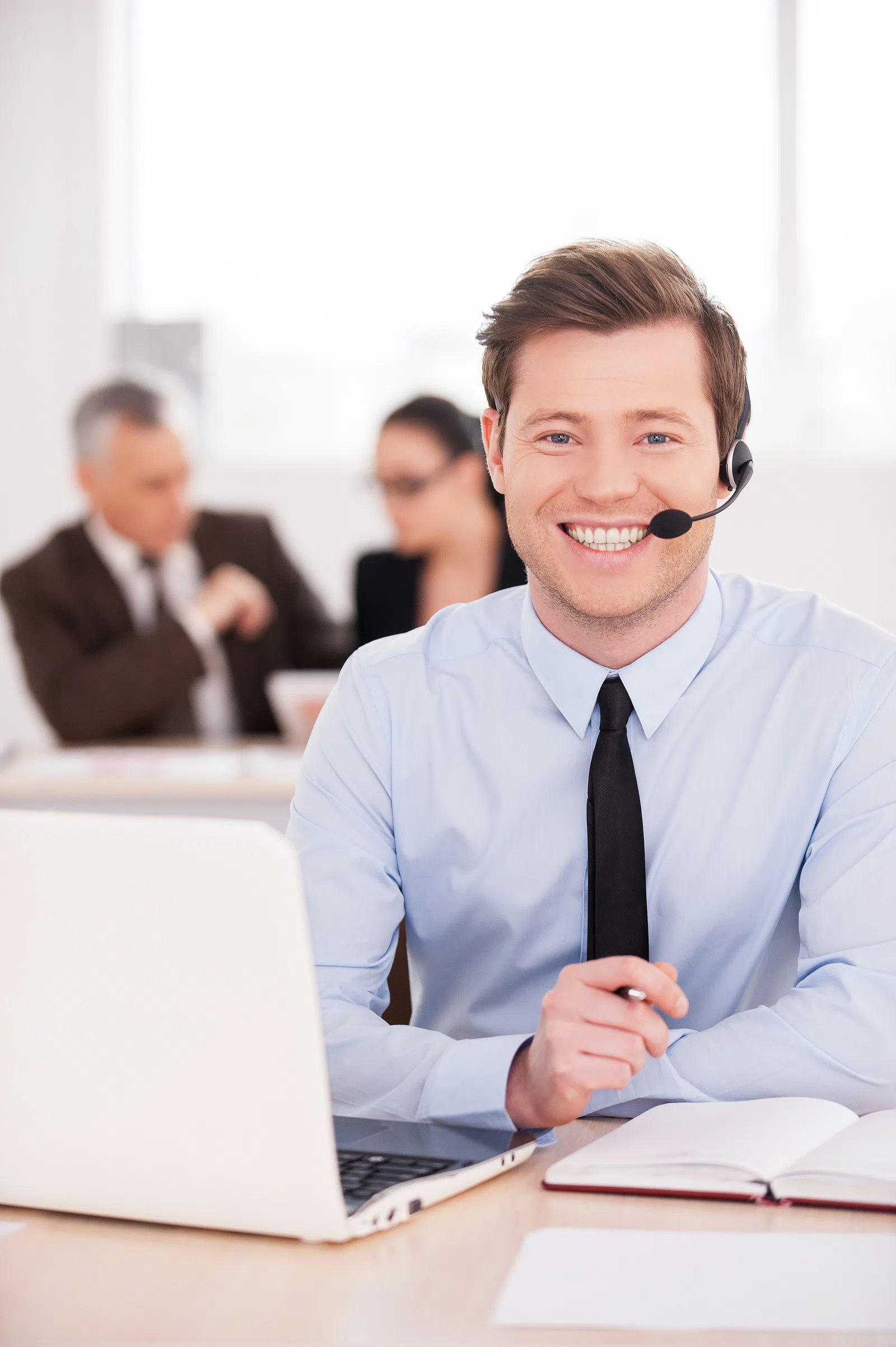 Smiling man in a headset at a desk with a laptop and notebook in a modern office environment.