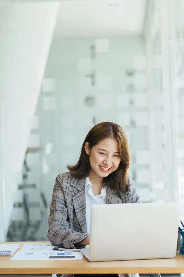 Young woman smiling while working on a laptop in a modern office environment.