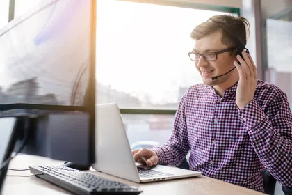 Man in checkered shirt working on laptop, using headset for customer support in bright office space.