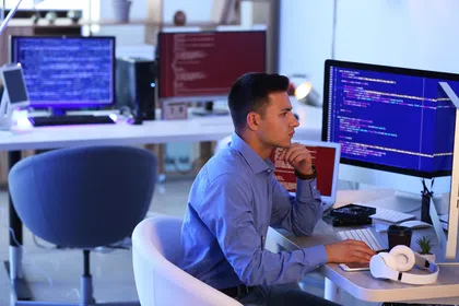 A focused man coding at a computer in a modern tech workspace with multiple screens.