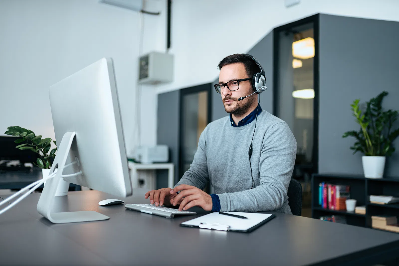 Man with glasses using a headset at a computer desk in a modern workspace.