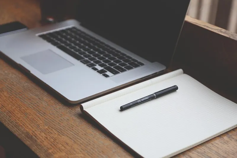 Open laptop beside blank grid notebook with pen on wooden desk workspace setup.