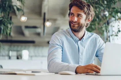 Smiling man working on a laptop in a modern office with plants and bright lighting.