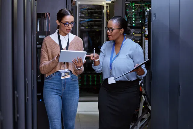 Two women discussing data in a server room, one using a tablet, the other holding a clipboard.