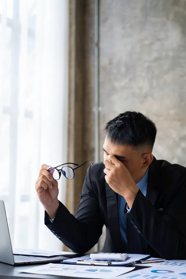 A stressed businessman adjusts his glasses while sitting at a desk with documents and a laptop.