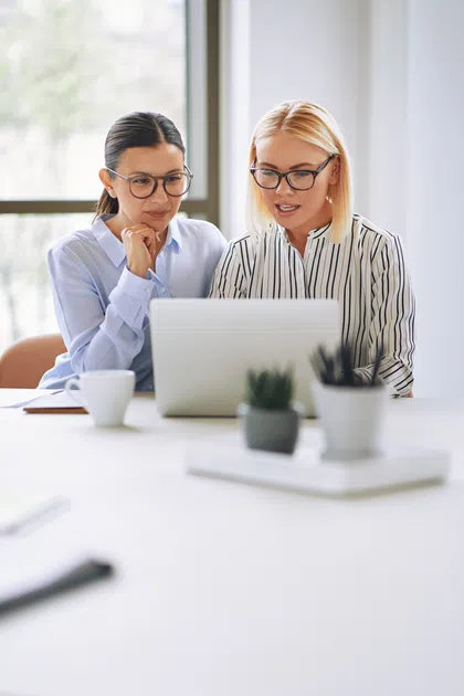 Two women collaborating while looking at a laptop in a modern office setting.