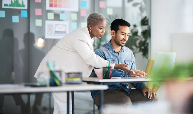 A businesswoman assisting a colleague with a laptop in a modern workspace.