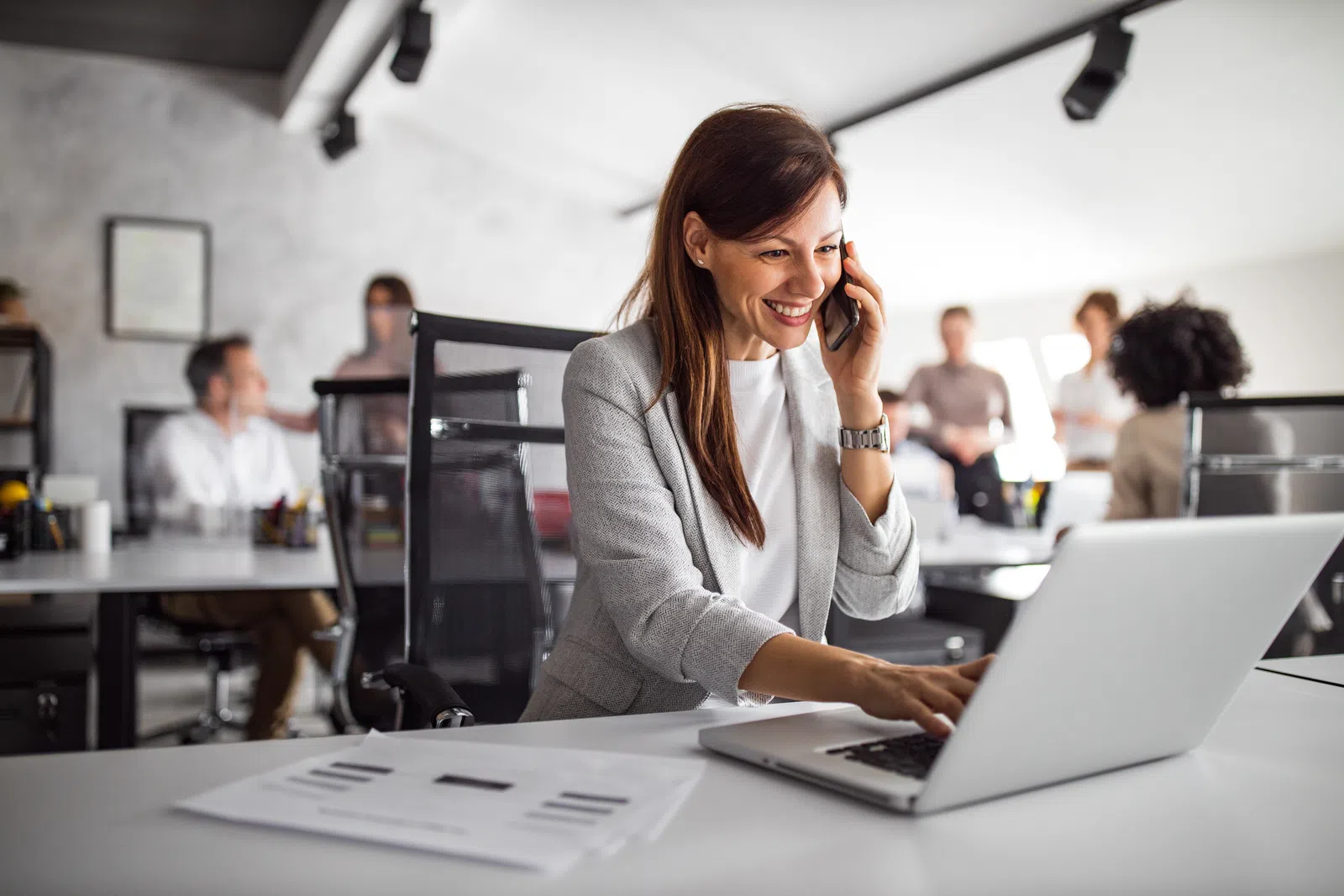 Smiling businesswoman on the phone working on a laptop in a modern office setting.