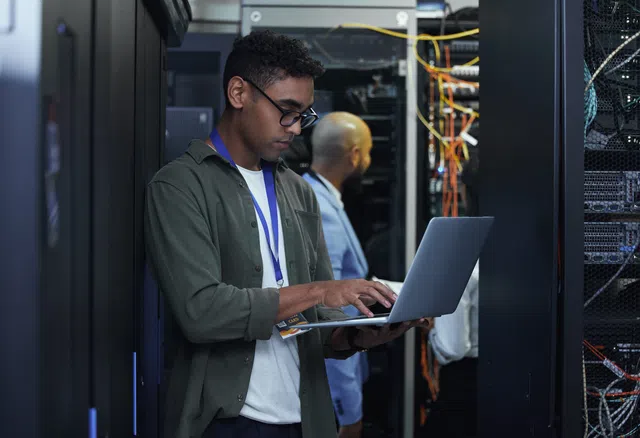 Man working on a laptop in a data center, surrounded by server equipment and wiring.