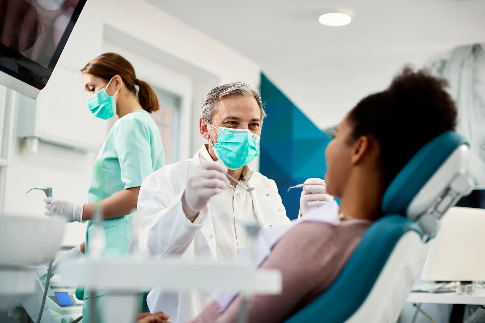 Dentist in mask consulting with patient in dental clinic, staff preparing in background.