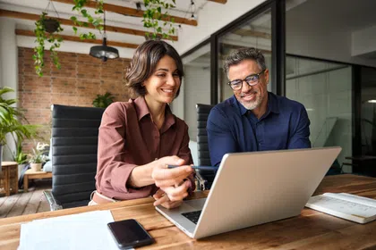 Two colleagues discussing work on a laptop in a modern office setting with plants.