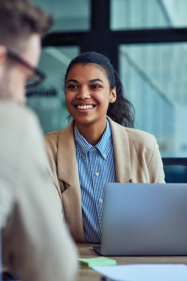 Smiling woman in professional attire engaging in conversation during a meeting with a laptop nearby.