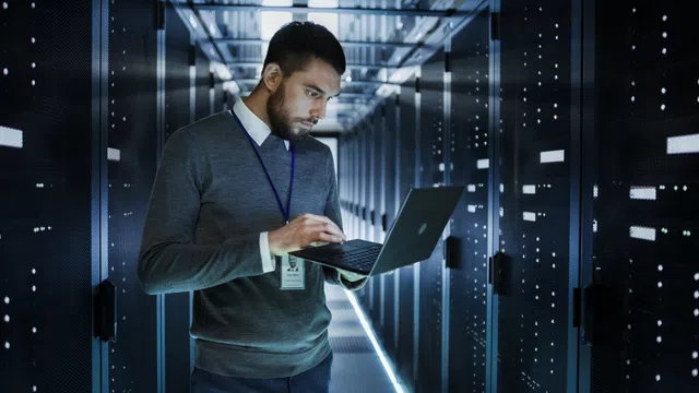 A professional working on a laptop inside a data center with server racks and bright LED lights.