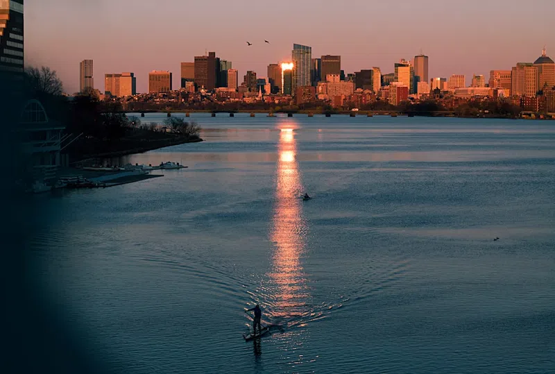 Paddleboarder on river at sunset with city skyline and reflective water under warm light.