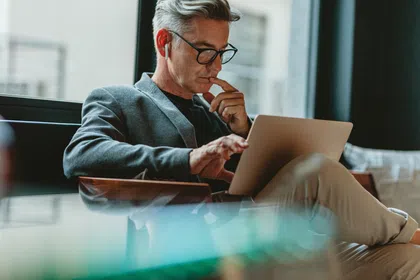 A thoughtful man in glasses using a laptop while sitting in a modern office lounge.