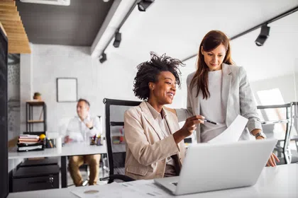 Two women collaborating at a modern office workspace with paperwork and laptops.
