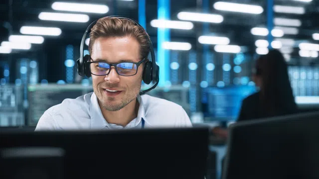 Man in headset smiling while working at a computer in a modern office environment.