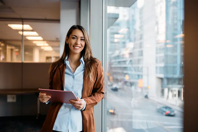 Smiling woman in a brown jacket holding a tablet, standing by an office window with a city view.
