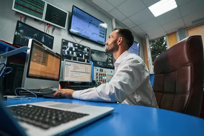 A man in a white shirt working at a control desk surrounded by multiple screens.