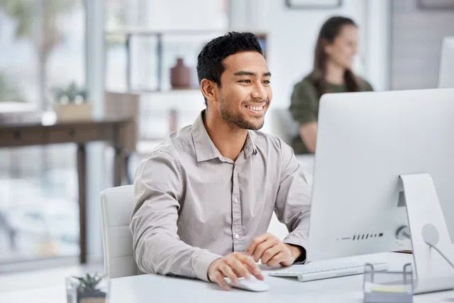 Smiling man working on a computer in a modern office setting with colleagues in the background.