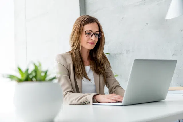 Professional woman working on a laptop in a modern office setting with a potted plant nearby.