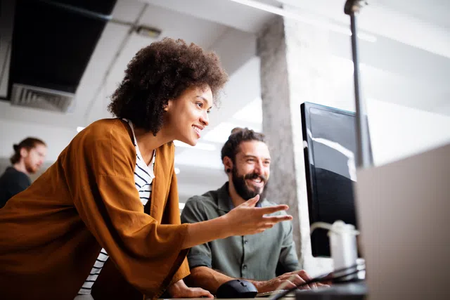 Two colleagues collaborating at a desk, one pointing at a computer screen in a bright office.