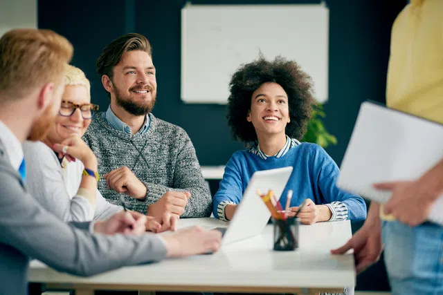 Diverse team in a meeting, smiling and engaged around a laptop and notepad on the table.