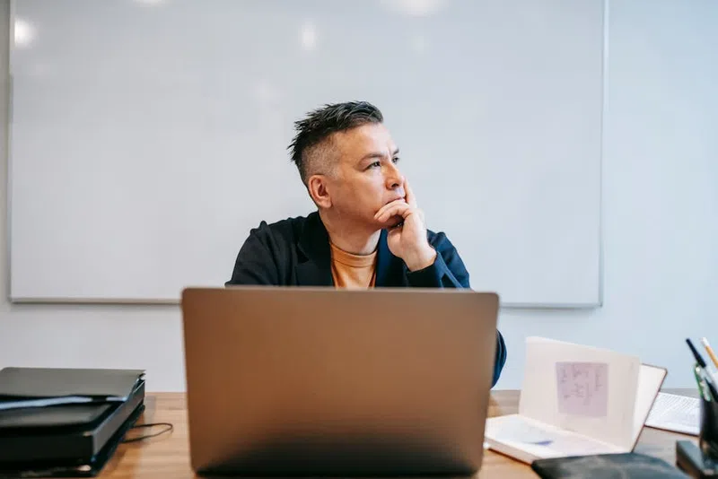 Man in casual jacket sitting at desk with laptop, looking thoughtfully to side in bright office space