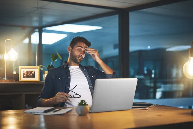 Frustrated man working late at desk with laptop, holding glasses in modern office setting.