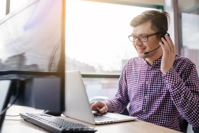 Man with headset working at a laptop, smiling in a bright office environment.