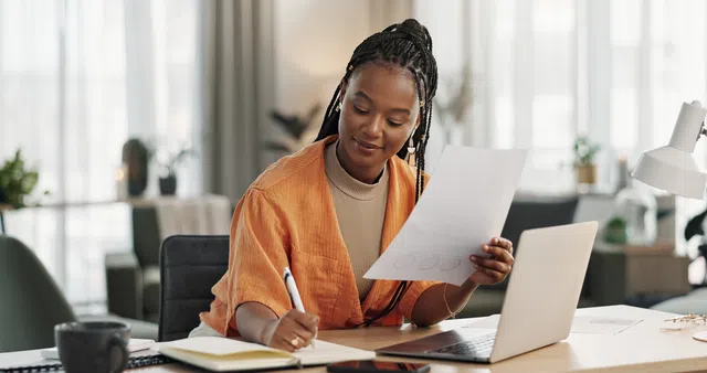 A woman working on a laptop, taking notes, in a bright, modern office space.