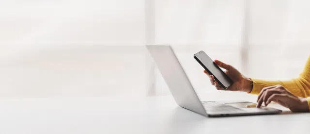 A person holding a smartphone while working on a laptop in a bright office setting.