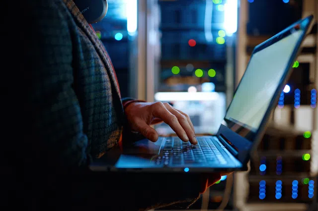 Person using a laptop in a server room with illuminated equipment in the background.