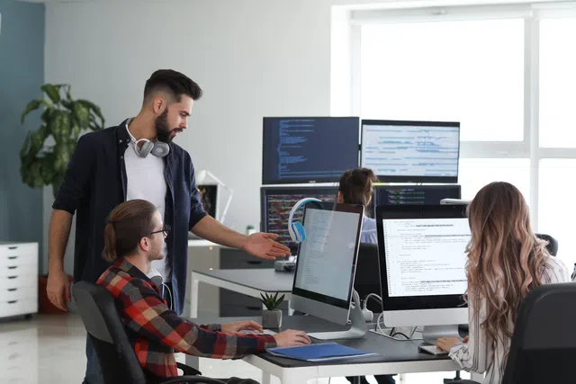 A team collaborating in an office, discussing work at computers with coding on screens and modern décor.
