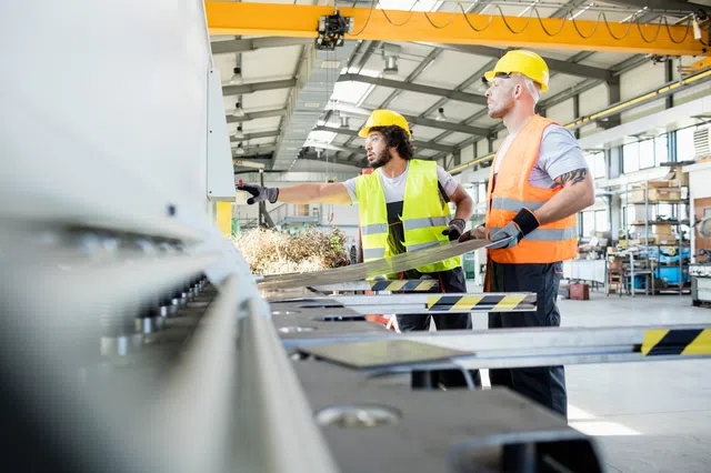 Two workers in hard hats and safety vests operating machinery in a bright industrial workshop.