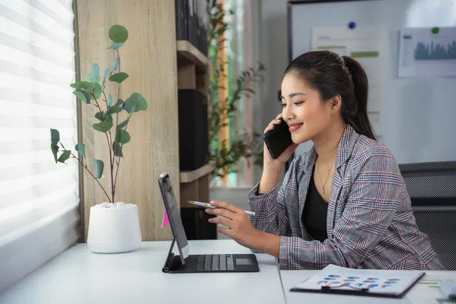 A professional woman working at her desk on a phone and tablet, with a plant in the background.