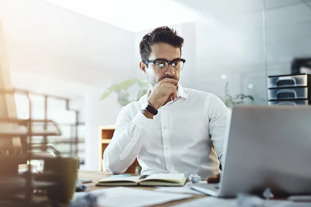Man in glasses working intently on a laptop in a bright office setting with notebooks and a coffee mug.