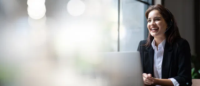 Smiling woman in a headset working on a laptop in a bright office environment.