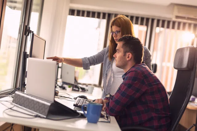 Woman and man collaborating at an office desk, focused on computer screen and discussing ideas.