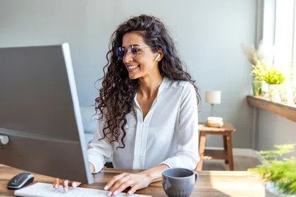 Smiling woman wearing glasses, working on a desktop computer in a bright home office.