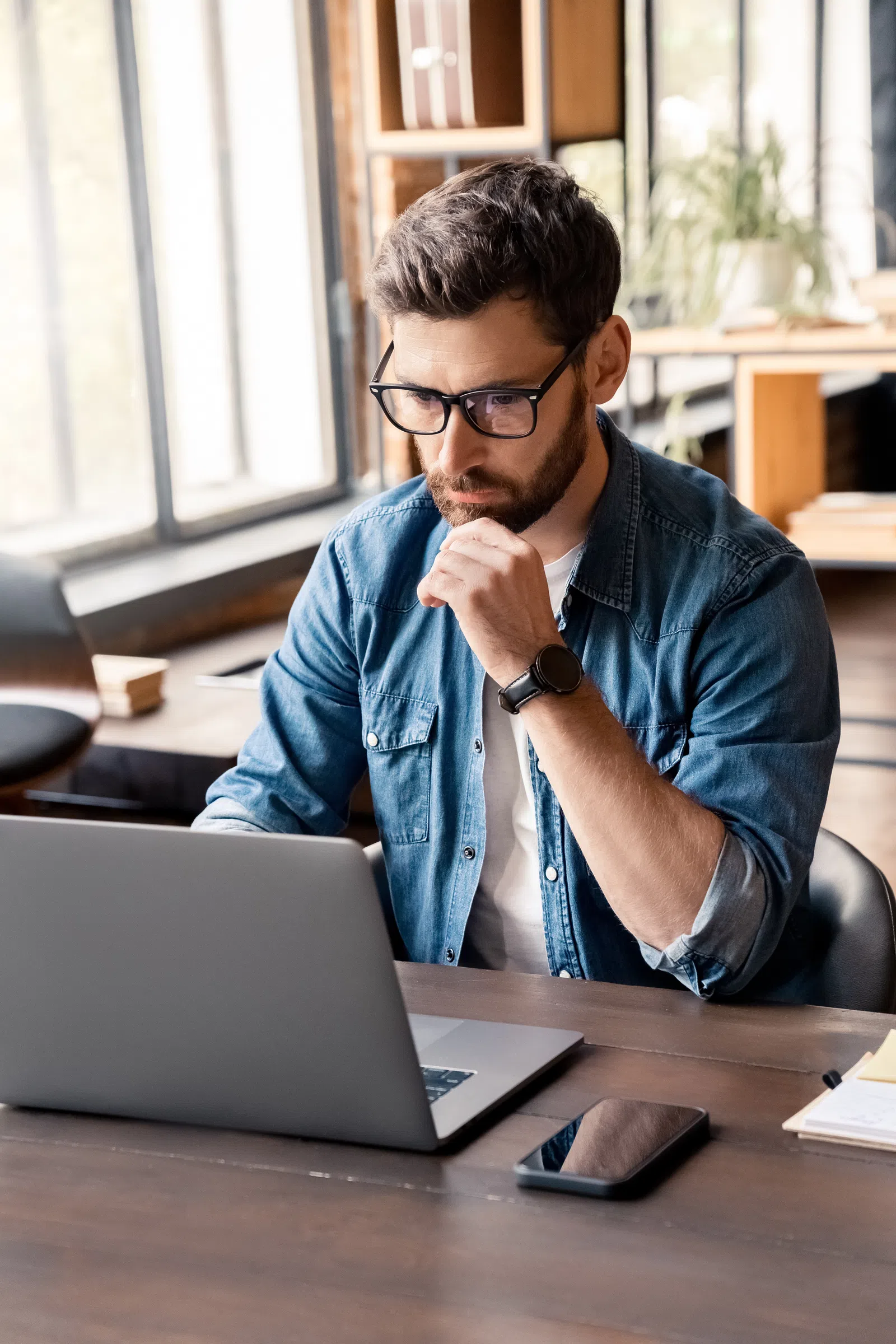 Young man with glasses working intently on a laptop in a modern office space.