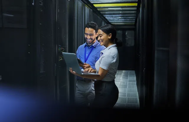 Two professionals collaborating in a data center, smiling while using a laptop and smartphone.