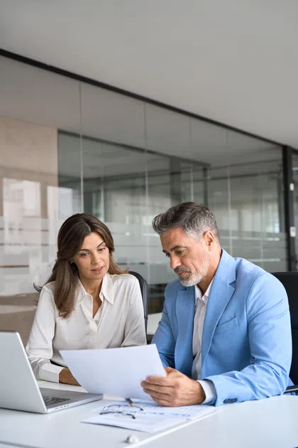 Two professionals discussing documents in a modern office setting with a laptop on the table.