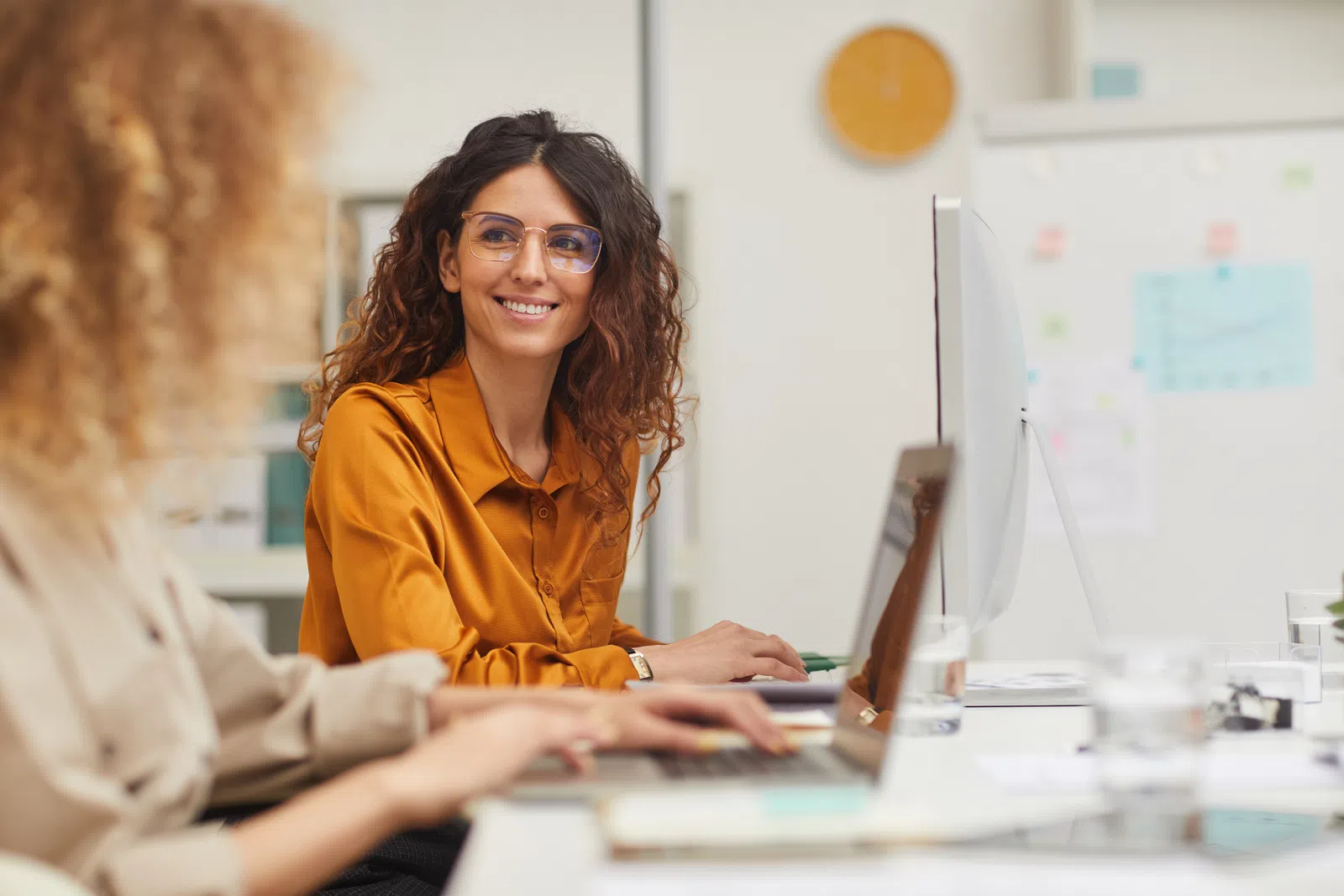 A woman in glasses smiles while working beside a colleague at a bright office desk.