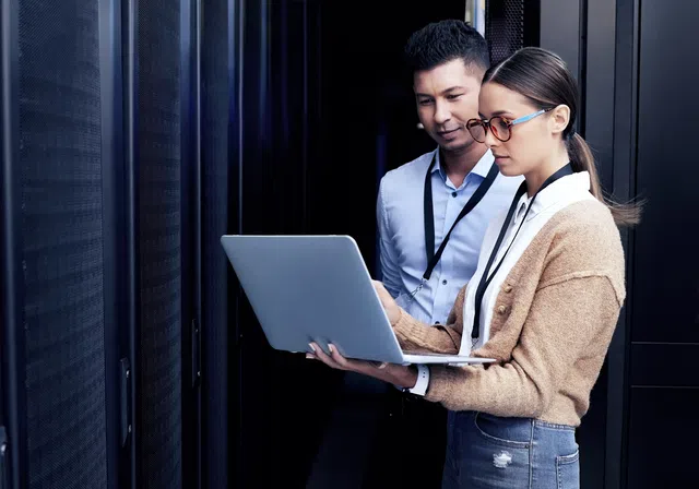 Two professionals collaborating on a laptop in a data center environment with server racks.