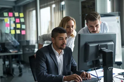 Professionals collaborating in an office, focusing on a computer screen with notes in the background.